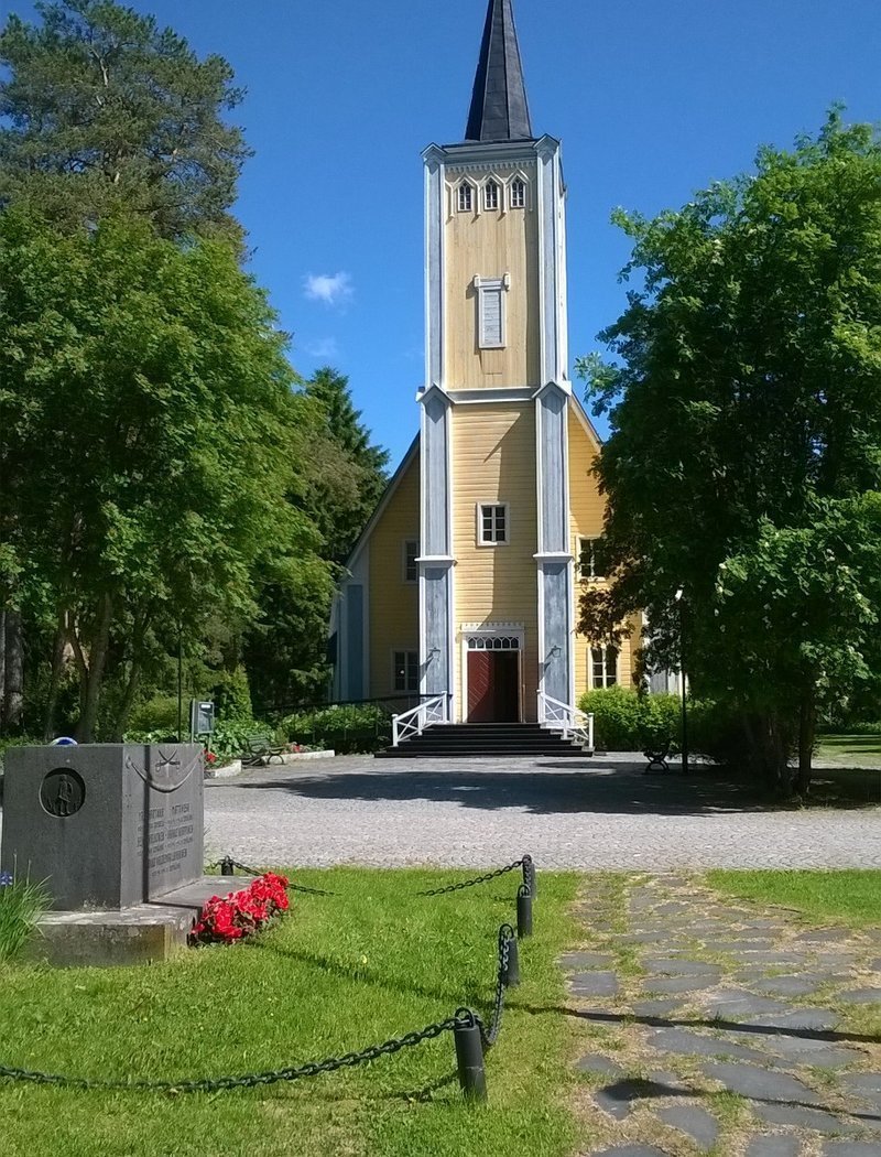 Kesäinen Muhoksen kirkko, kivilaattakäytävä, muistomerkki rajattu kettingillä, punaiset kukat muistomerkin edessä